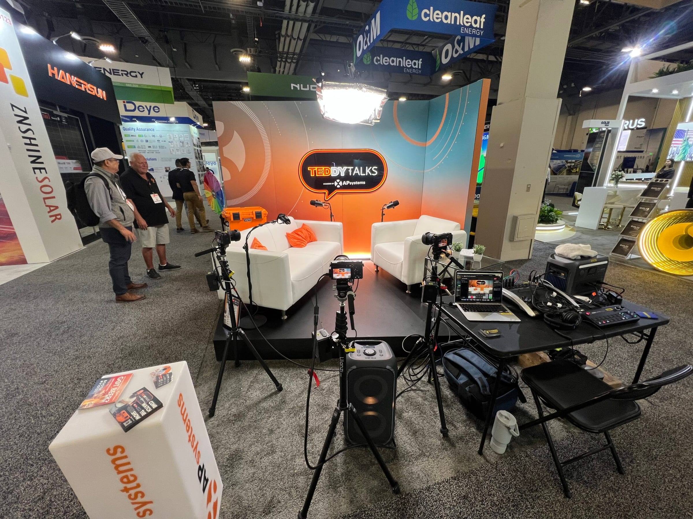 The Teddy Talks podcast booth for AP Systems at RE+ in Las Vegas, with two white sofas, cameras on tripods and lights on the show floor