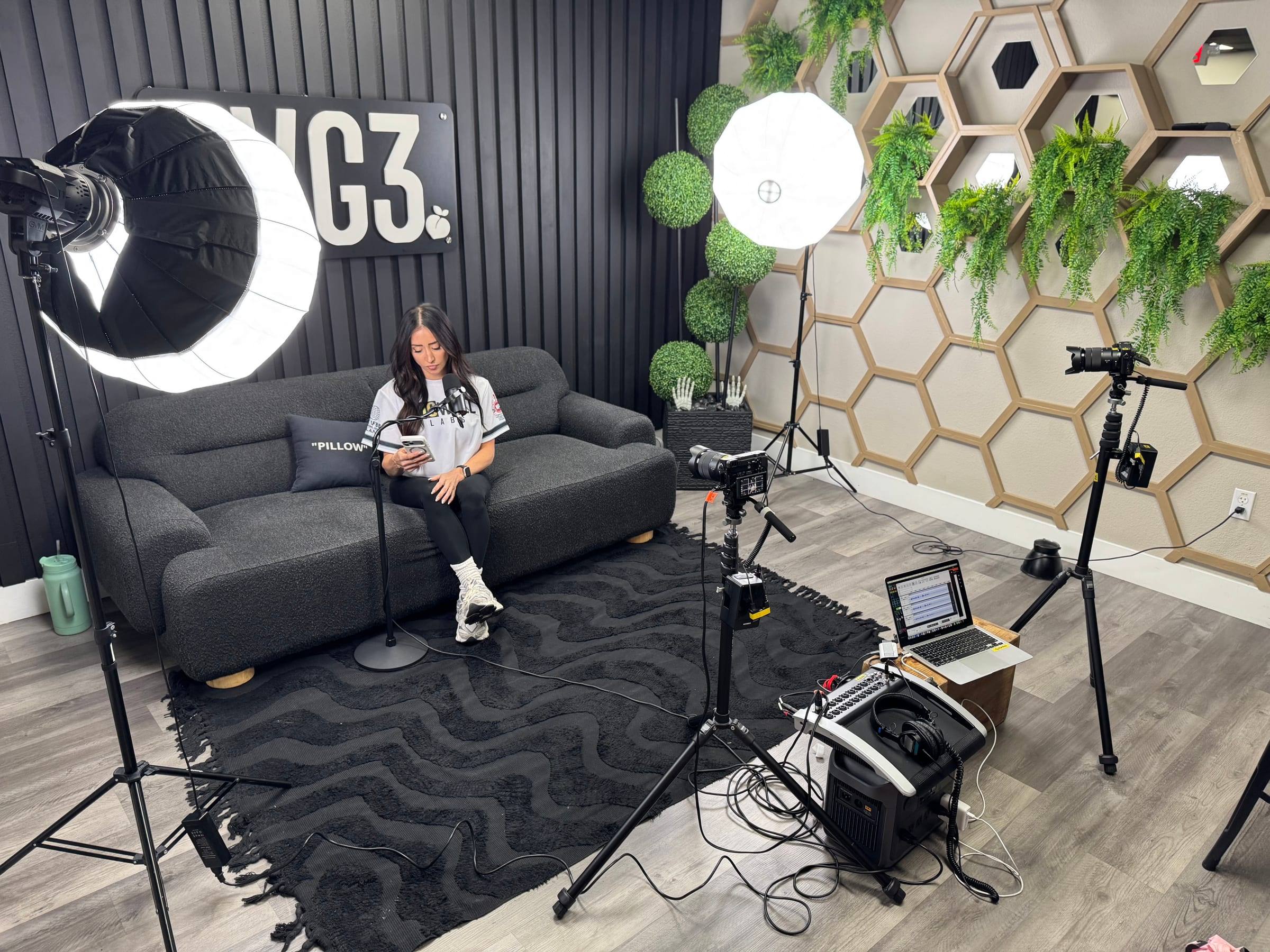 An office lounge set up as a podcast studio with lantern lights, cameras and a host on a gray sofa